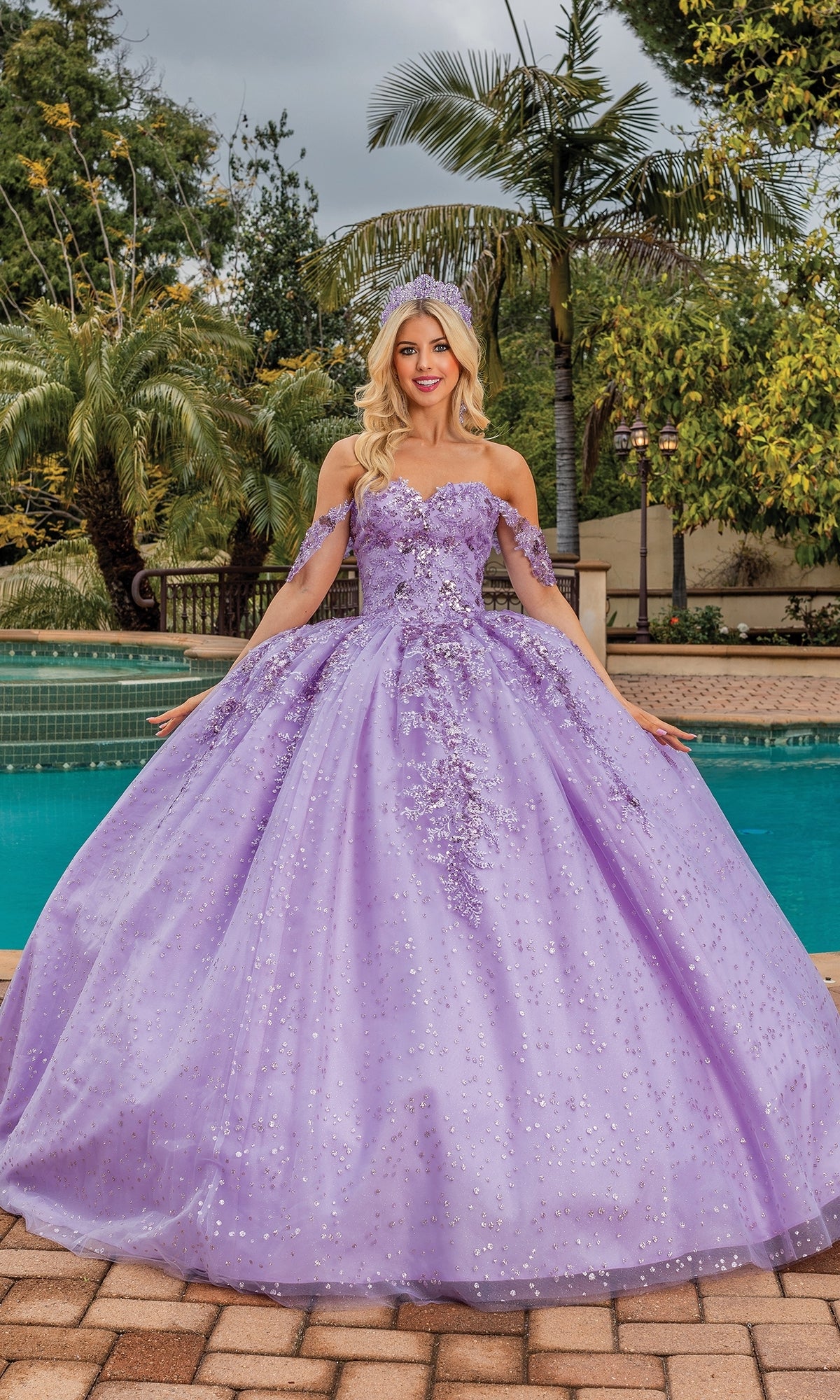 A woman stands by a pool surrounded by palm trees, wearing the Quinceanera Dress 1862 By Dancing Queen—an off-the-shoulder ball gown with floral embellishments and a sweetheart neckline.
